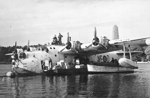 Fliegendes Boot auf einem See, mit Männern, die Kisten in ein Boot entladen.