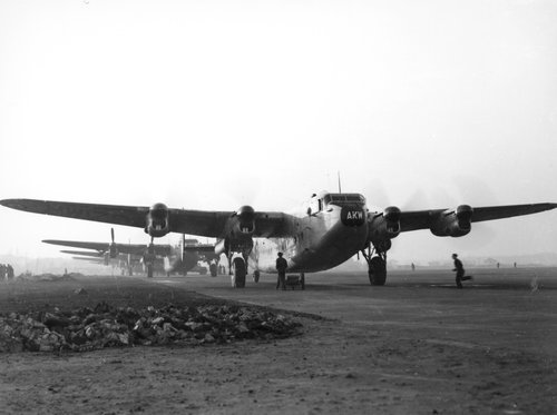 Eine Reihe viermotoriger Transportflugzeuge mit laufenden Motoren bilden eine Schlange auf einem Flugfeld.